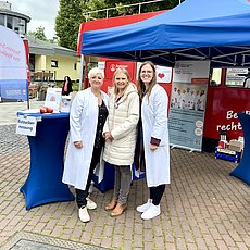 Drei Frauen in weißen Arztkitteln vor einem blauen Pavillion mit Informationsstand