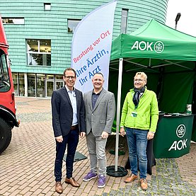 Drei Männer (zwei Ärzte in Anzügen, ein Vertreter der Krankenkasse) vor einem grünen Pavillion der AOK 